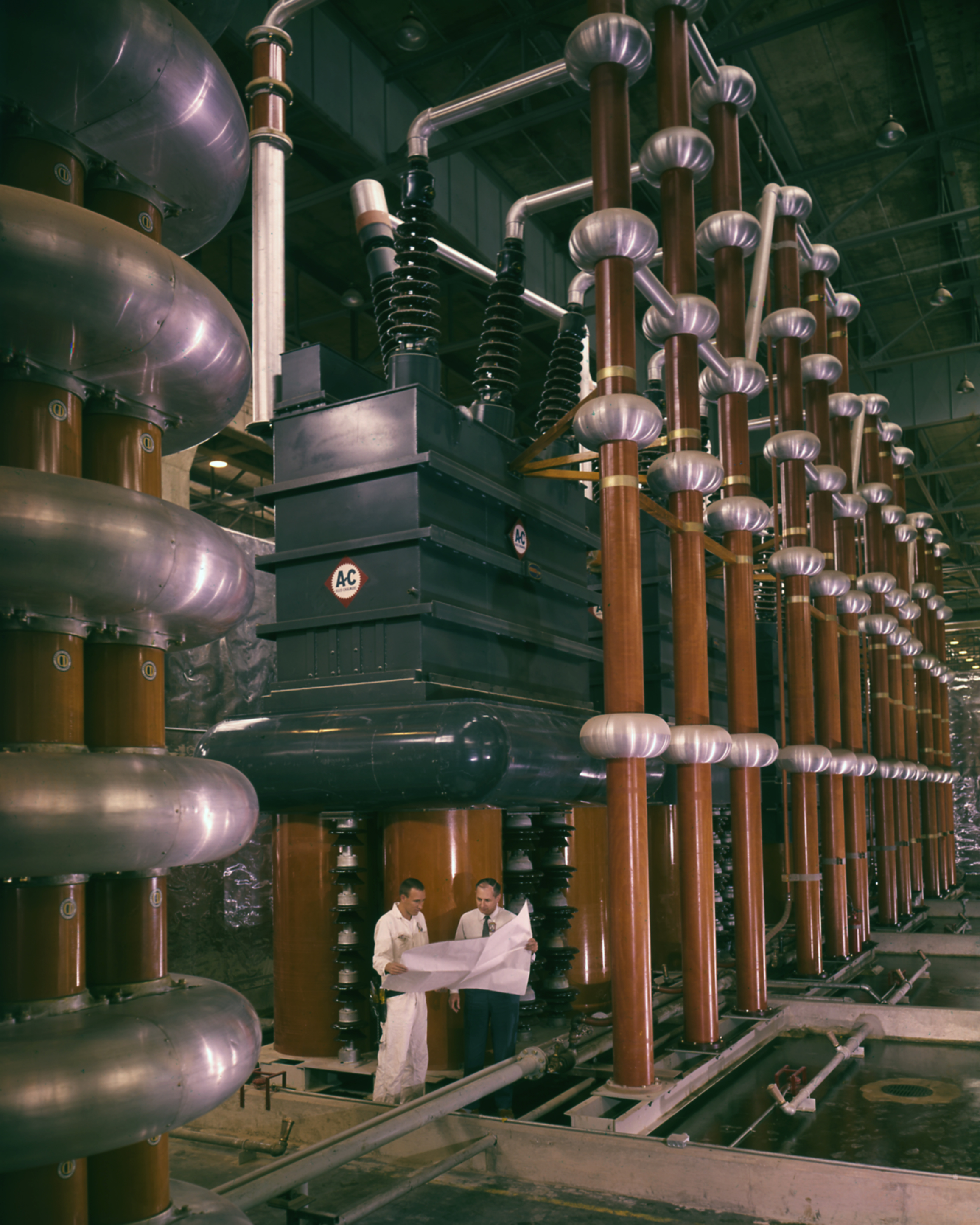 The electrical infrastructure for the DCX in Geneva. Rows of transformers and an electrical box, with two engineers standing underneath holding paper blueprints.