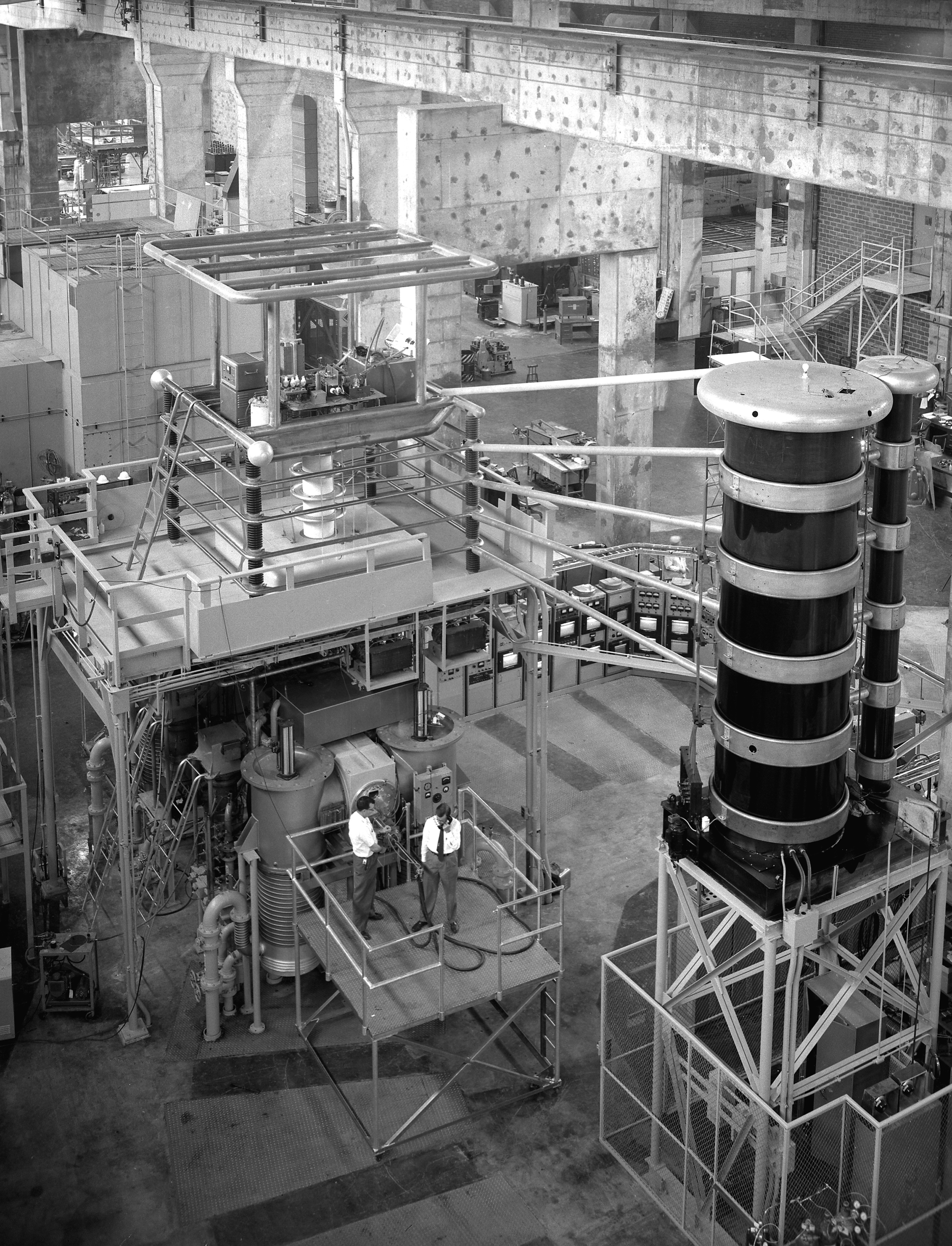 The Direct Current Experiment at ORNL, depicting two men standing on a platform, dwarfed by a large black and silver coil tower, with lots of pipes and conduits running all around them.