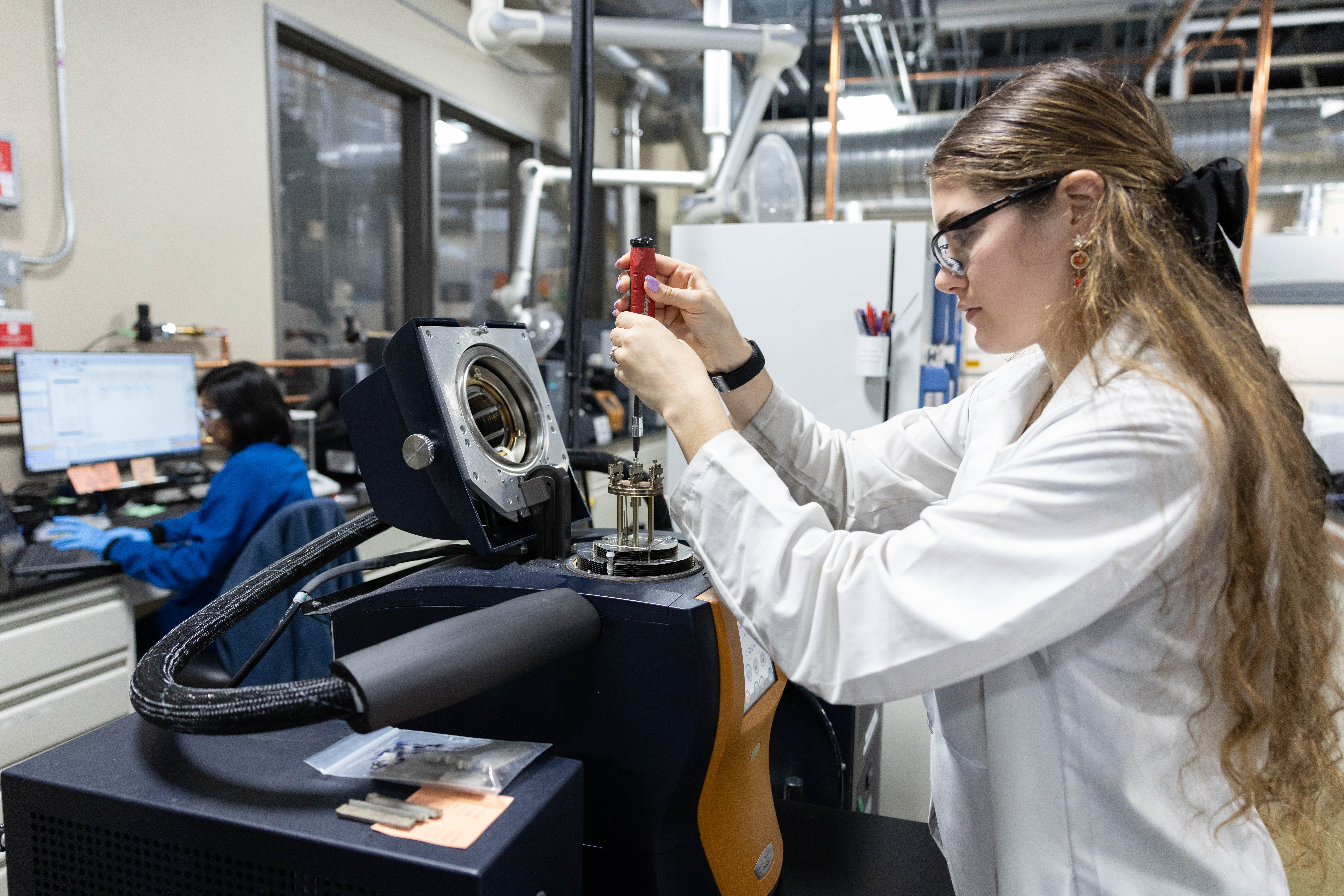 A researcher in a white lab coat and safety glasses uses a precision tool to adjust equipment in a materials science lab, while another scientist works at a nearby computer workstation in the background.