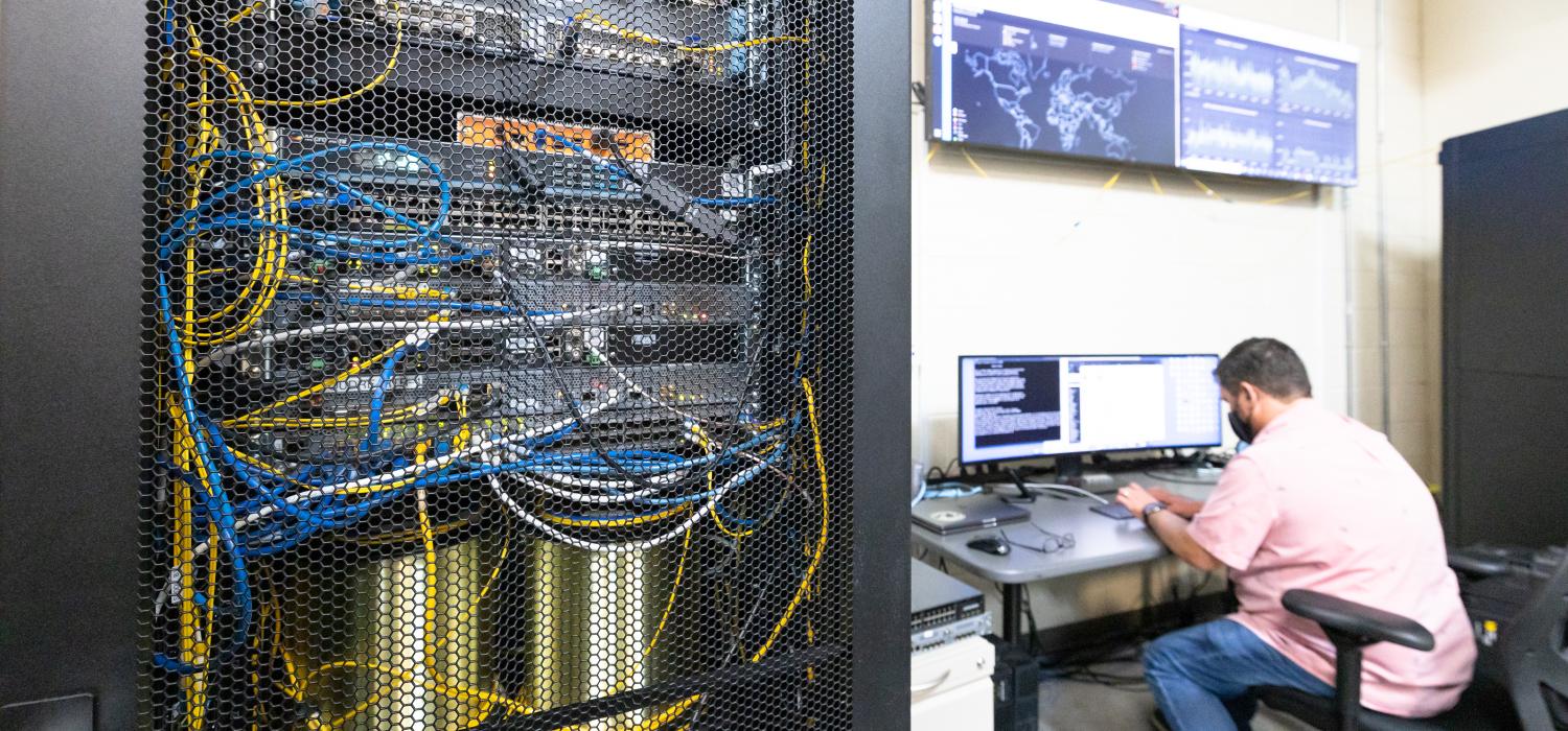 Electronics case with visible woring showing, man with back to camera working at desk in background beneath screens showing maps and graphs