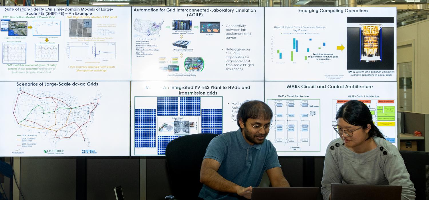 A man and woman researcher look at information on their laptops with a large screen behind them showing research slides