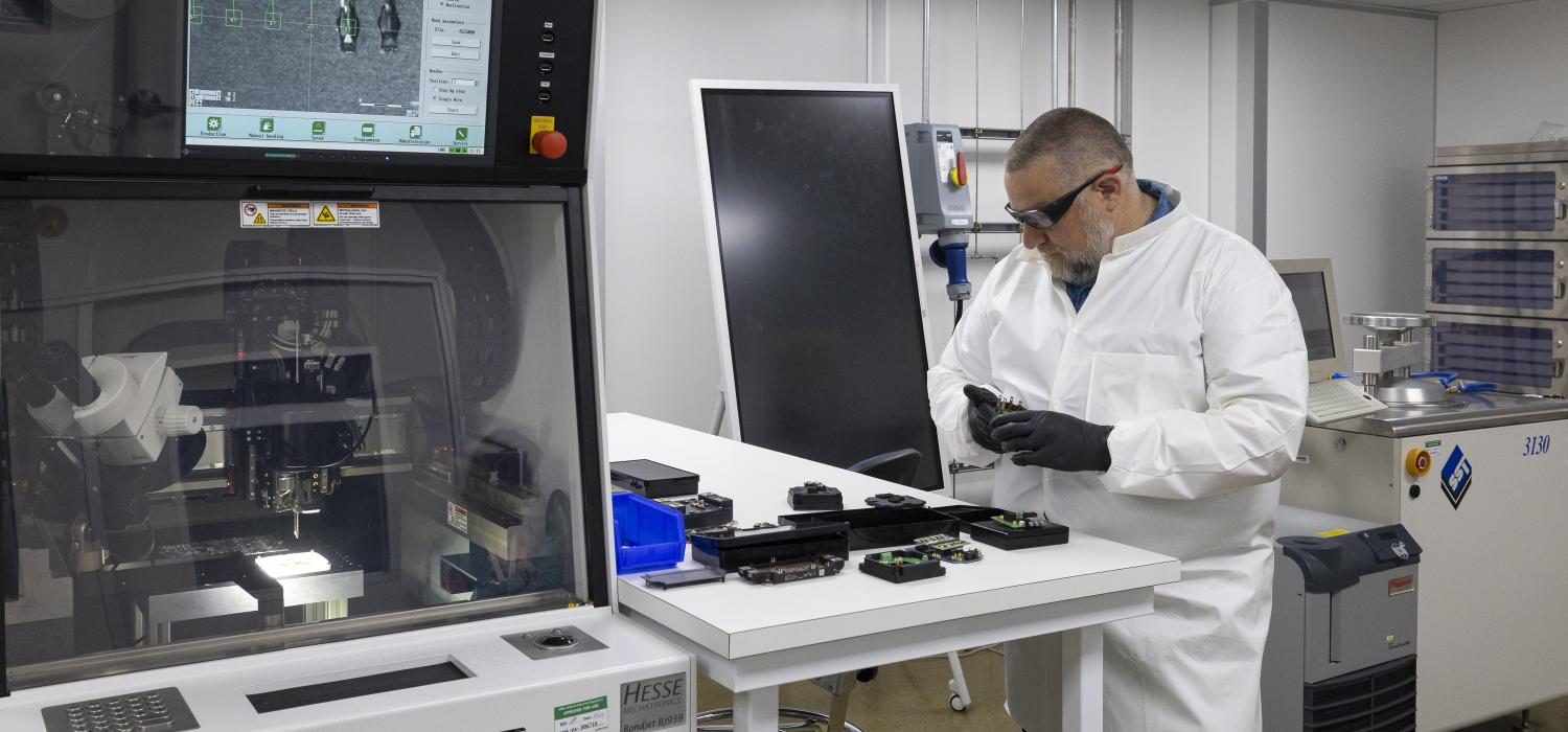 Researcher in a white lab coat, gloves and safety glasses works on power modules next to a wire bonder machine in a lab