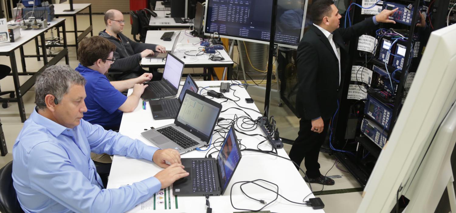 Three men sitting at laptops and one standing punching buttons in electrical equipment inside tall metal cabinets in a lab
