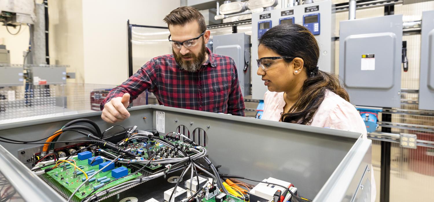 A man and woman in a lab stand over a metal cabinet filled with power electronics connected by cords