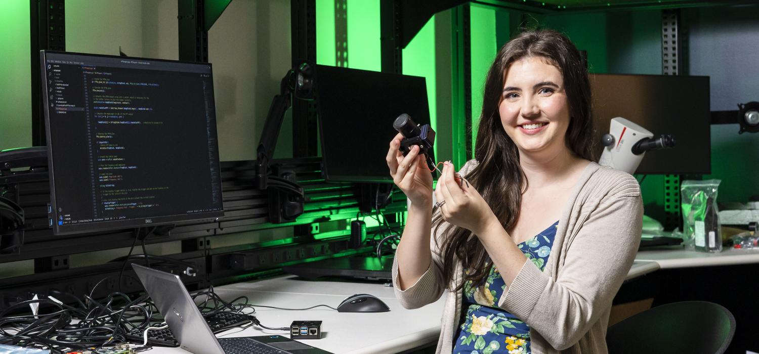 A woman working in a cyber laboratory smiles at the camera while holding research equipment