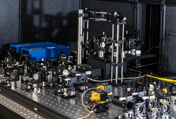 A complex optical laboratory setup featuring an array of precision-mounted mirrors, lenses, and optical components on a perforated metal table. A structured metal frame supports additional optical devices, while blue electronic equipment is positioned in the background. Yellow fiber optic cables run along the surface, indicating connections for data transmission or laser experiments.
