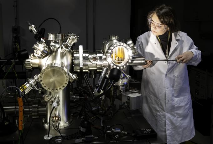 A researcher wearing a white lab coat and safety glasses works with a large stainless steel vacuum chamber in a laboratory. She uses a tool to manipulate an experiment inside the illuminated chamber, which features multiple ports, wiring, and scientific instruments. The setting includes advanced scientific equipment used for materials or quantum research.