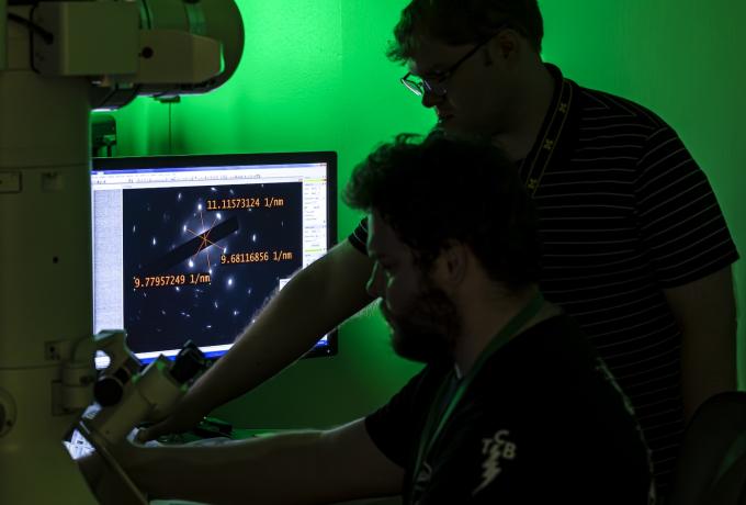 Two researchers operate a high-resolution electron microscope in a darkened lab with green lighting, analyzing atomic-scale diffraction data displayed on a monitor.