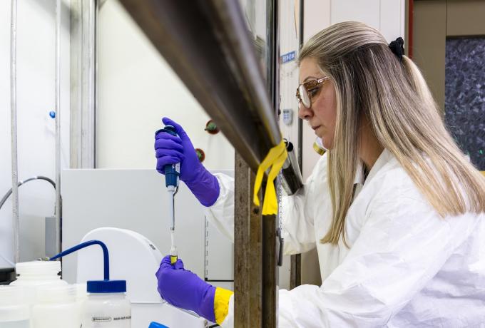 A researcher wearing safety glasses and protective clothing uses a pipette to transfer liquid into a vial inside a laboratory fume hood, demonstrating careful chemical handling.