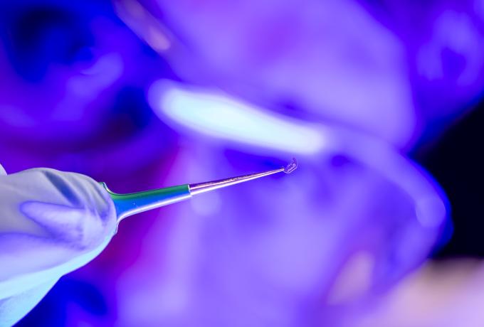 A gloved hand holds precision tweezers gripping a tiny, transparent microdevice under bright purple-blue lab lighting, highlighting advanced materials or microfabrication research.