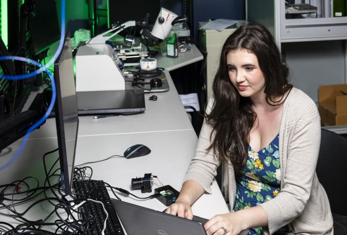 A woman types on a computer with microscopes and computer equipment in the background