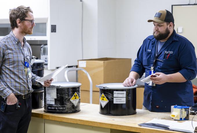 Two people stand next to a table with black cannisters filled with actinium-225.