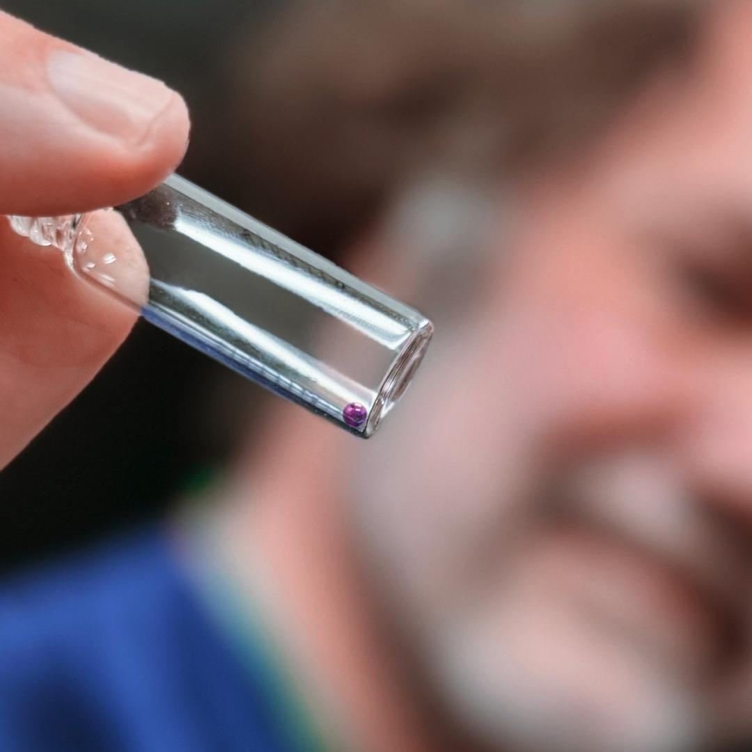 Man in a beard holding tweezers, showing a bead if space glass closer to the screen.  