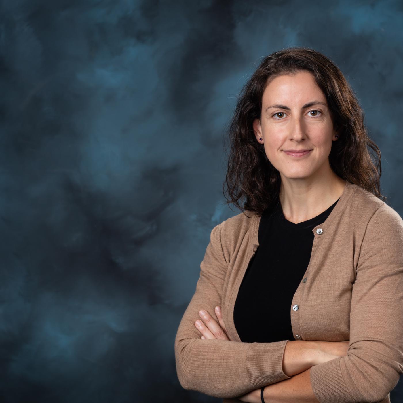 Image is of woman with dark hair crossing her arms and posting for a photo in front of a blue backdrop. She is wearing a black shirt with a tan cardigan. 