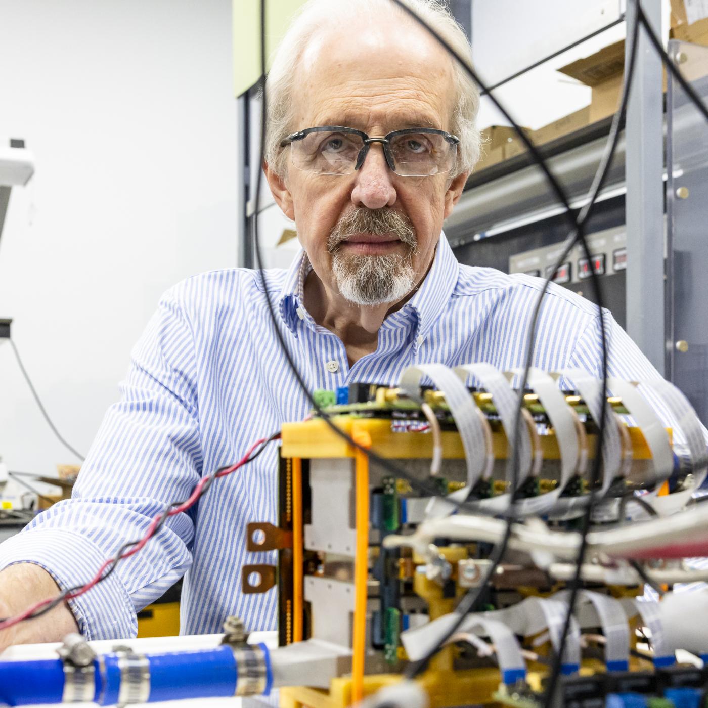 Scientist is in his lab with wires and electronics in the foreground, one with a medium yellow box. 