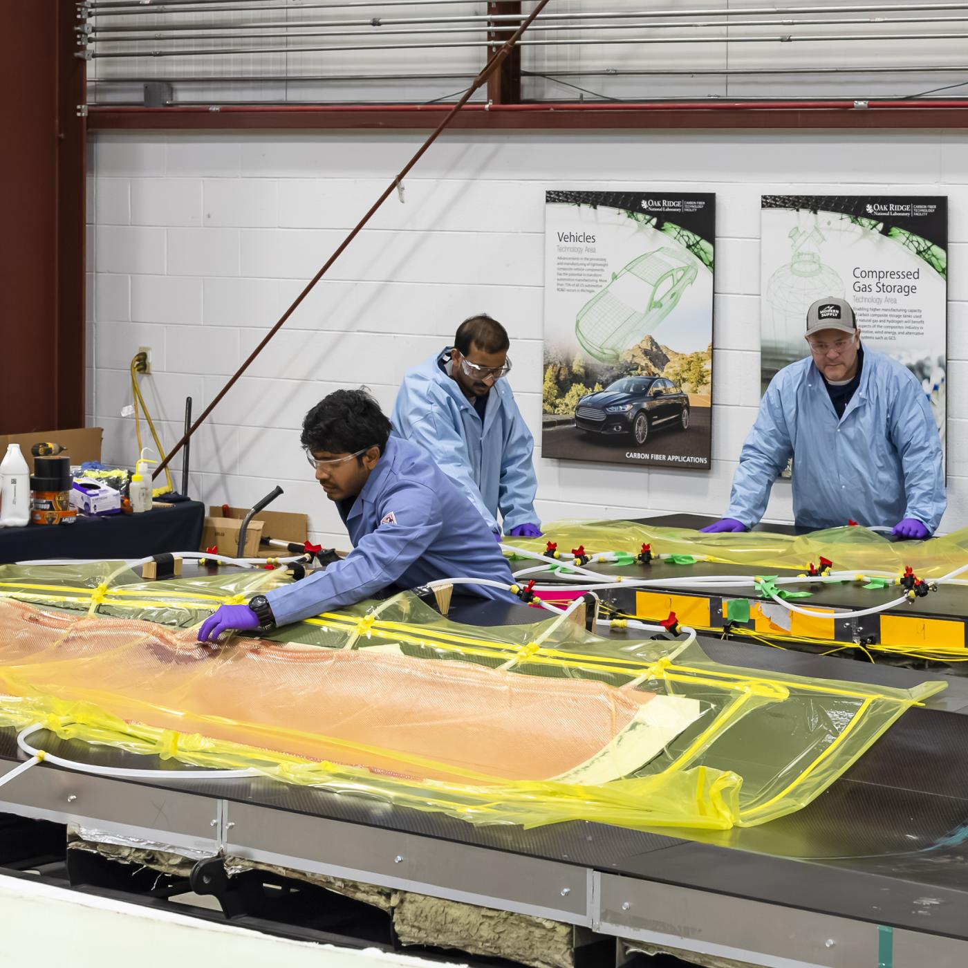 5 scientists in blue and white coats are leaning over the wind blades covered in orange and yellow material