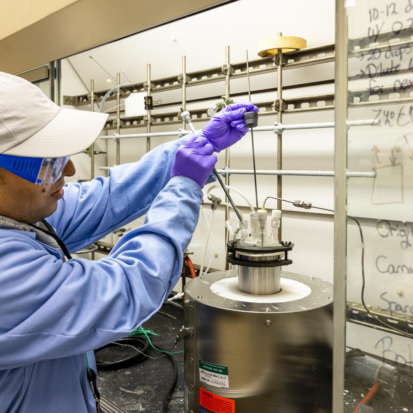 Researcher in a blue coat and glasses, purple gloves and white baseball gat pulls out materials from a metal canister