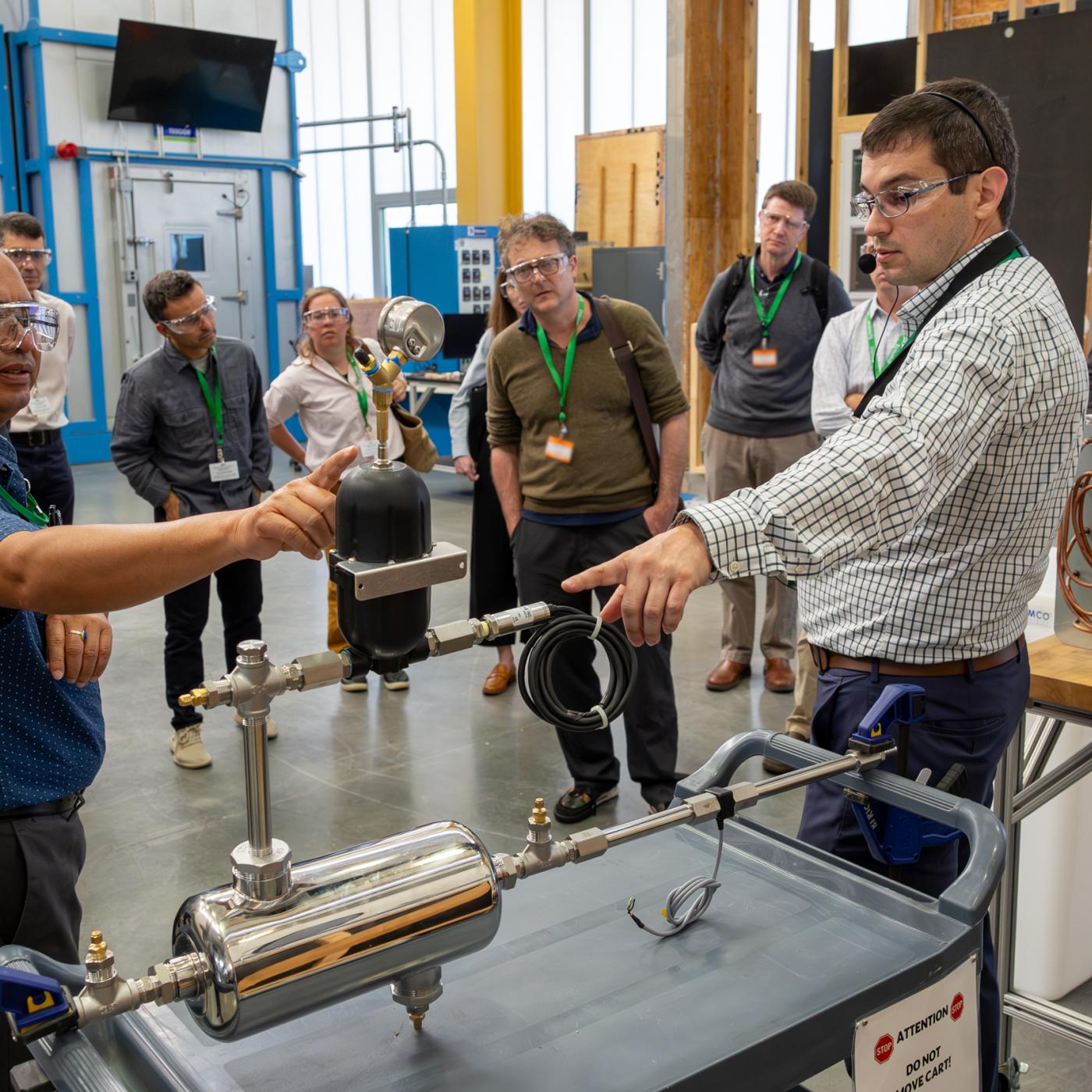 Two scientists are standing in the lab pointing at an object on the table, both wearing glasses. 