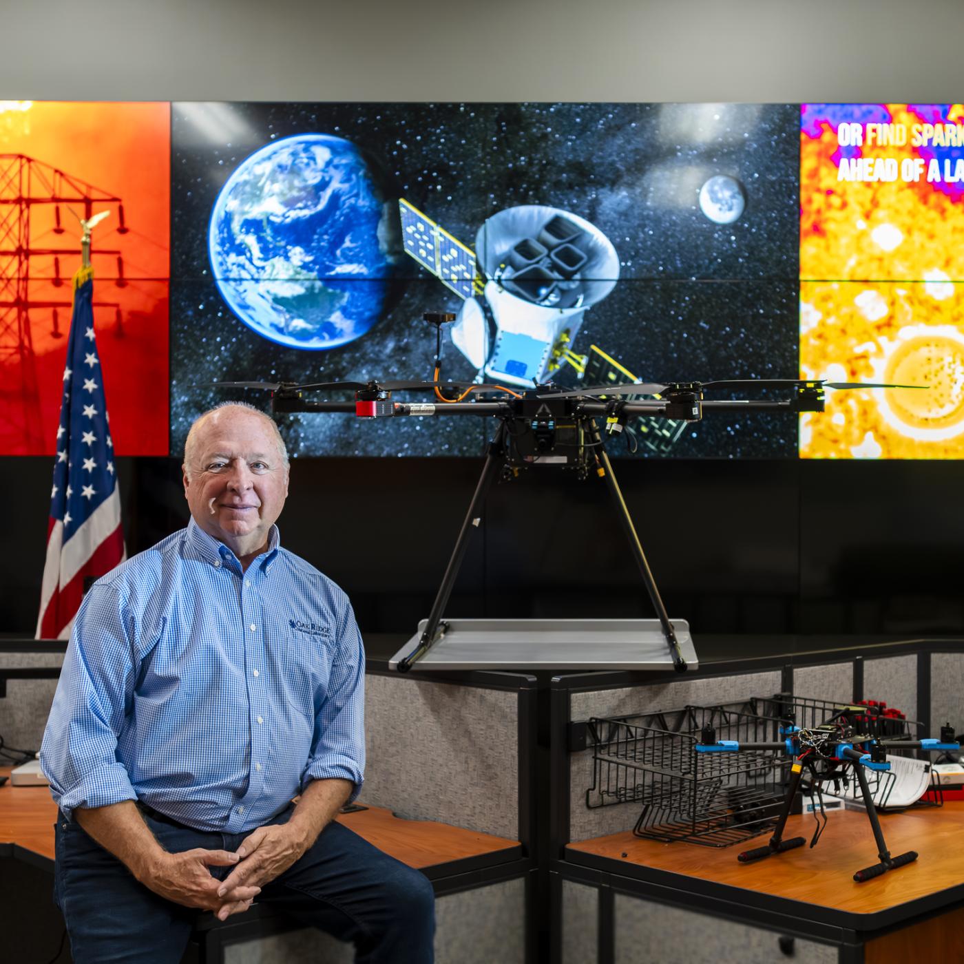 ORNL researcher is sitting on a desk with his hands crossed, three screens behind him depicting work in satellites and space. Pictures on the left and right are orange in color while the middle photo is blue and reflects an image from space