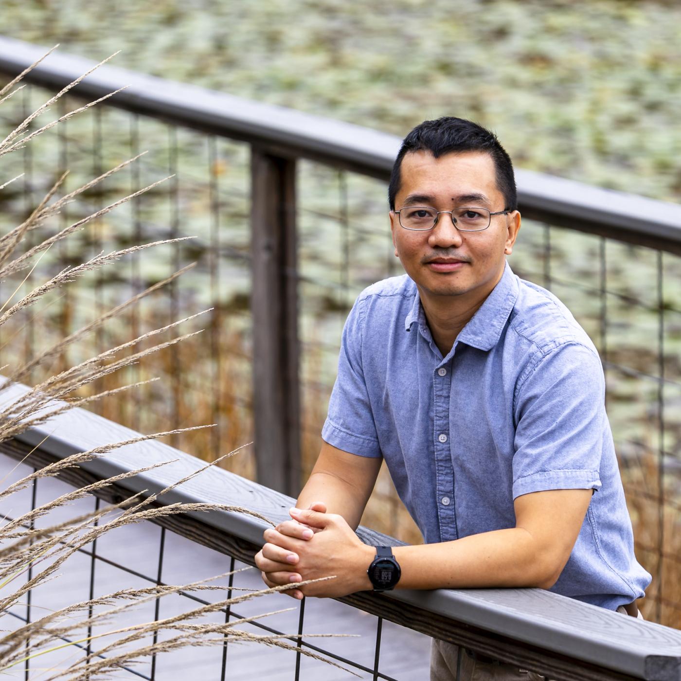 ORNL researcher Phong Le poses for a photo outside on a walkway bridge over the pond. The photo is framed with brown and green plants