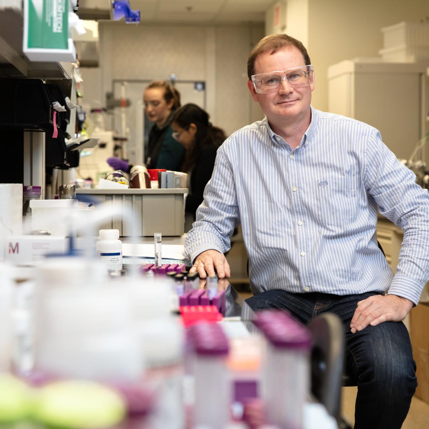 Hugh O'Neil, director or ORNL's Center for Structural Molecular Biology is sitting in the lab on a stool, hand on desk with glasses on. There are lab related items blurred in the foreground. 