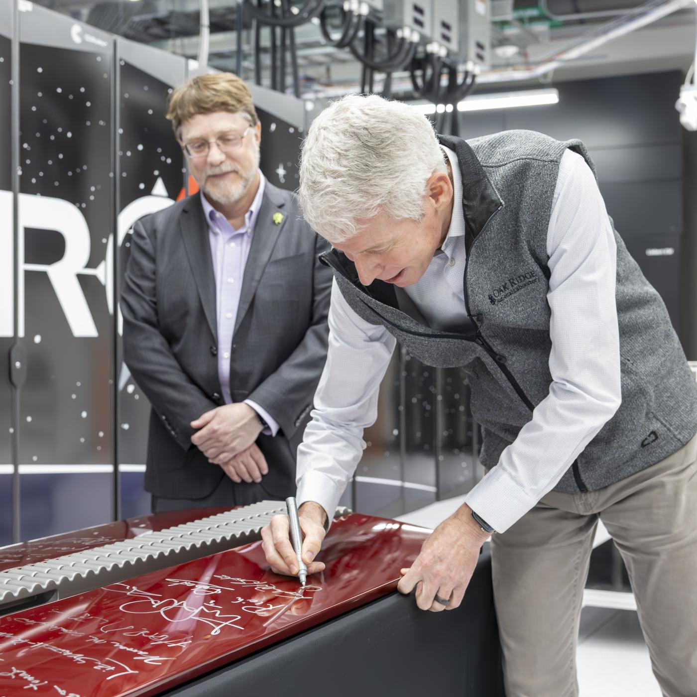 Secretary Wright leans over red computer door, signing with silver sharpie as ORNL Director Stephen Streiffer looks on