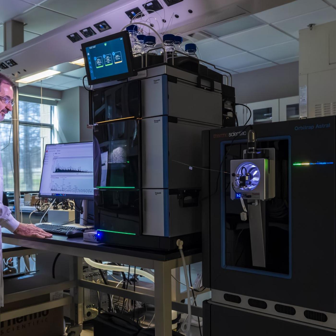 Scientist standing beside mass spectrometry equipment in a laboratory, with instrumentation panels and analysis tools visible in the background