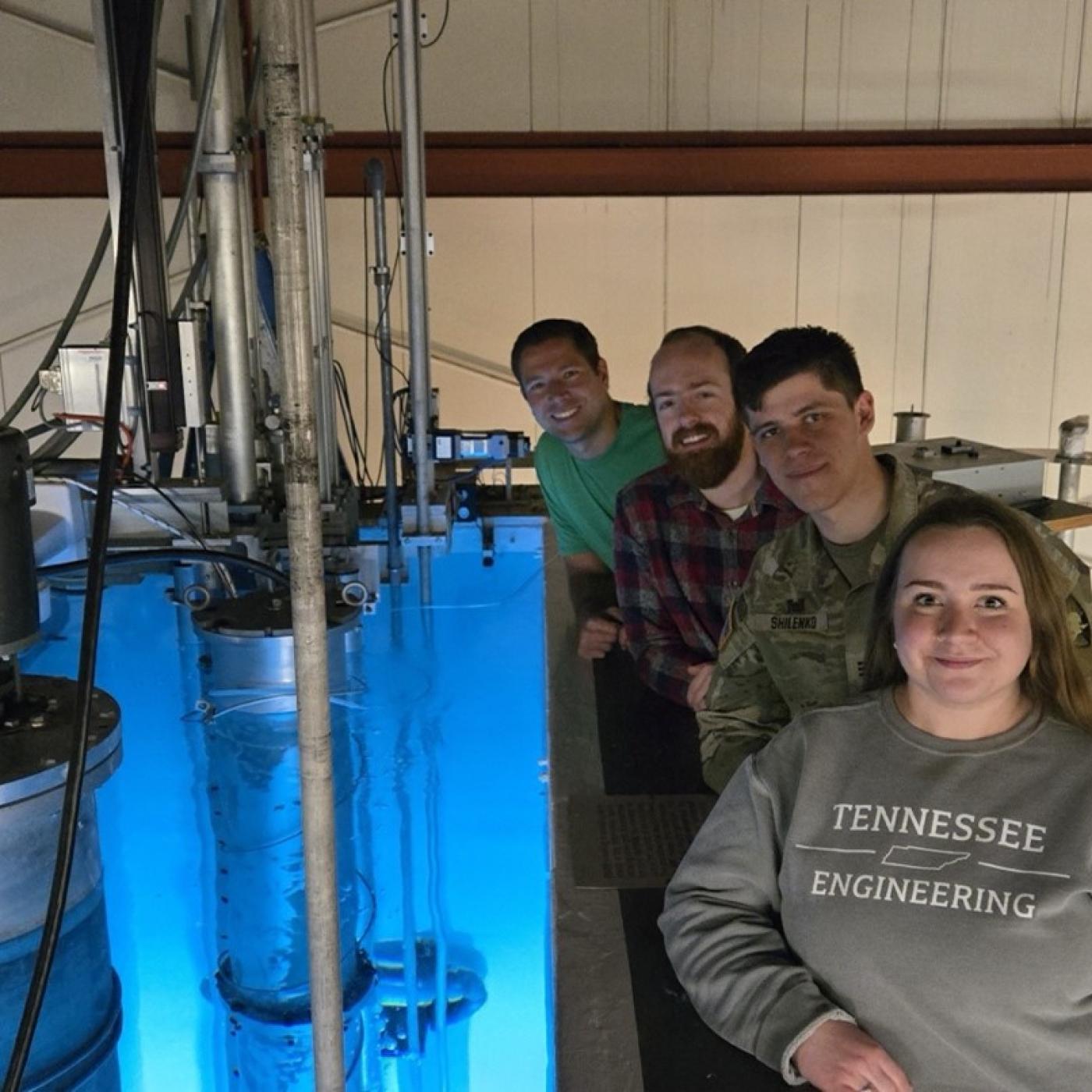 Four researchers are standing next to a research rector that is glowing blue 