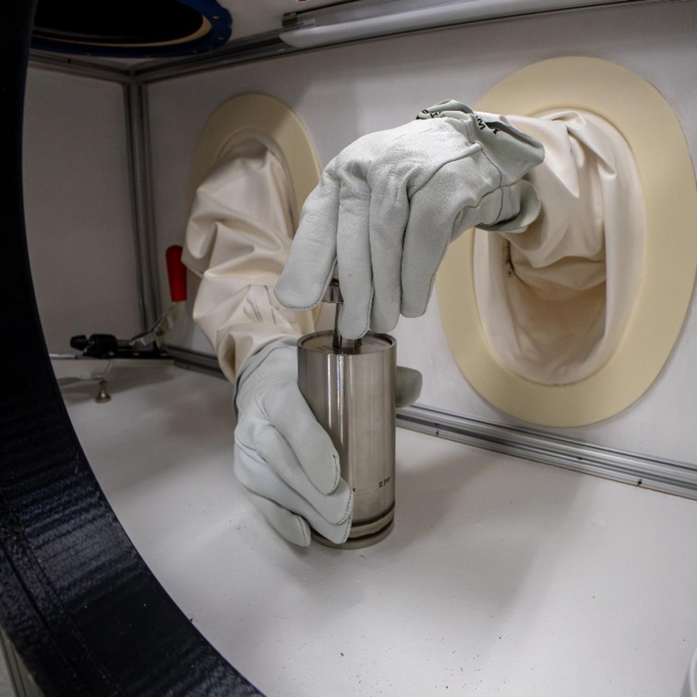 Two hands in gloves are holding a silver tube inside a white box with openings for the hands to poke through
