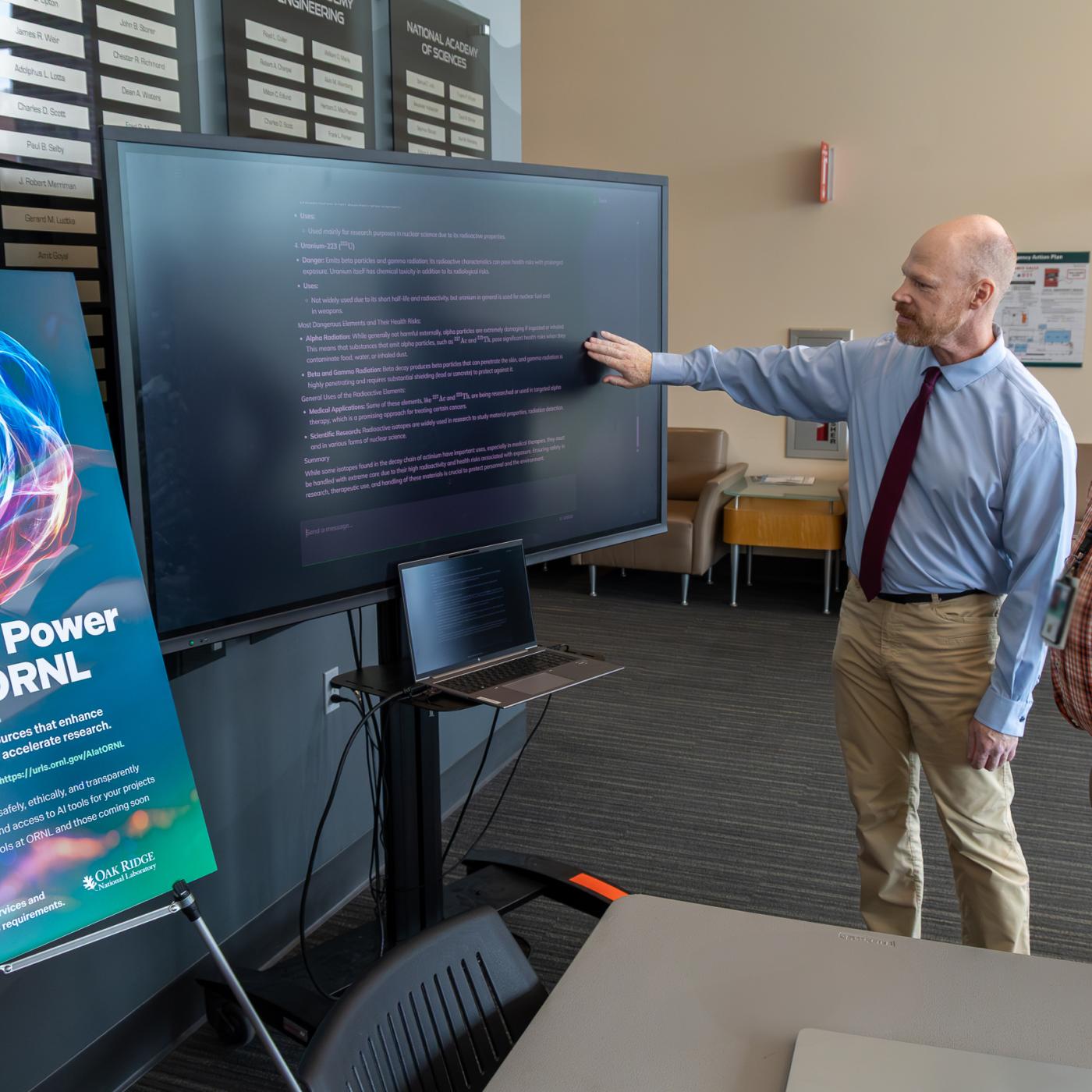 Two ORNL researchers are standing to the right of a computer screen and a poster promoting the AI Expo