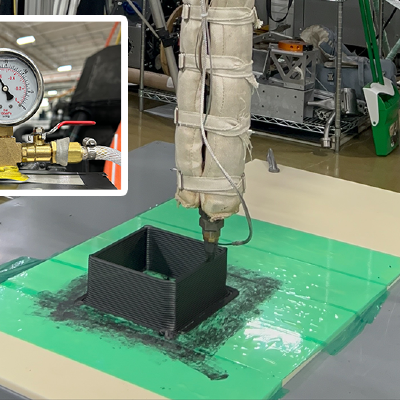 A 3D printing nozzle wrapped in insulation extrudes black composite material into a small square mold on a green and white flat surface in a lab setting. Inset shows a close-up of a pressure gauge connected to brass valves and tubing.