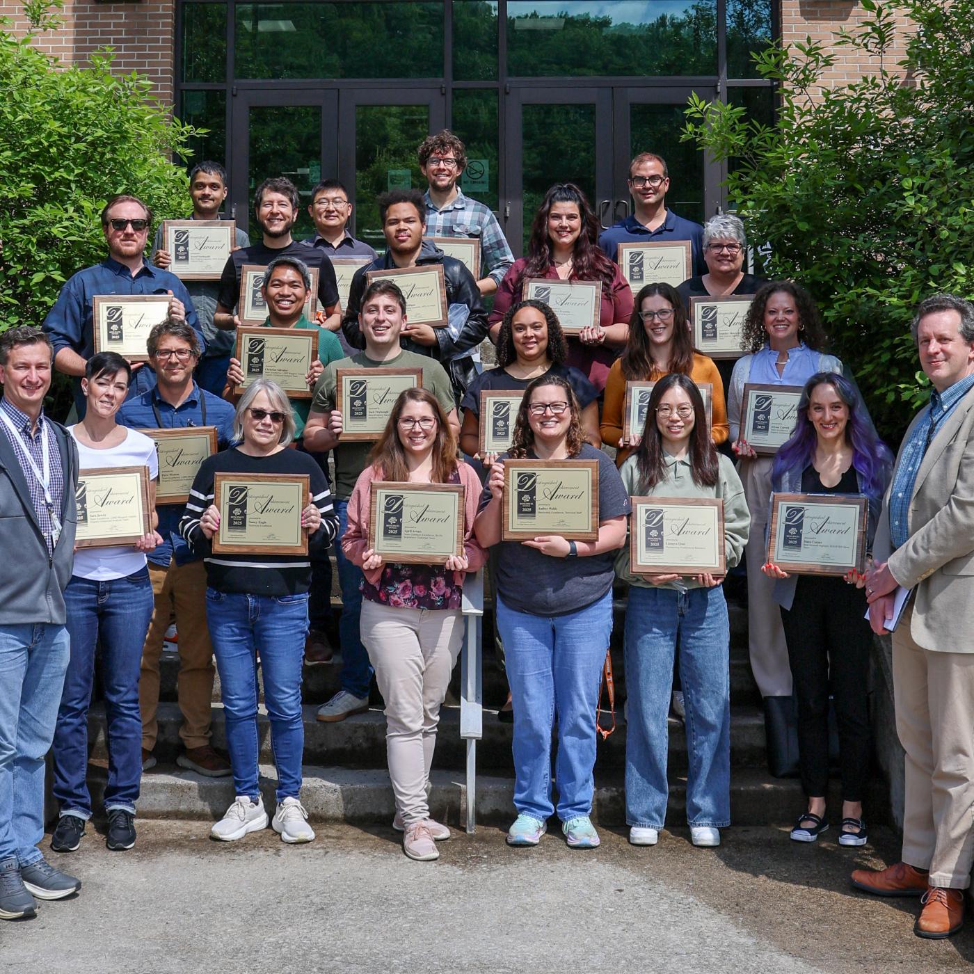group photo taken in front of a brick building of people holding award plaques