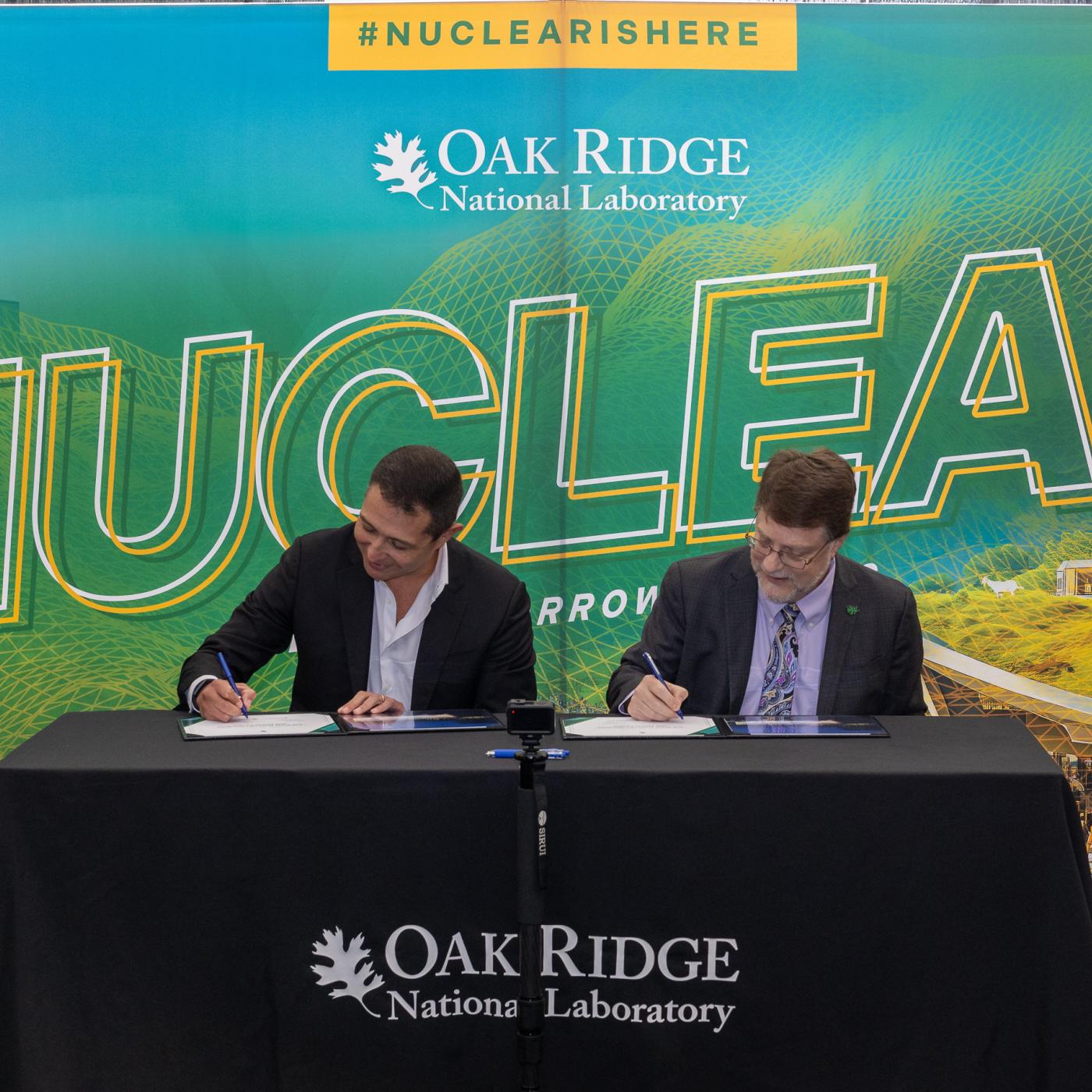 MOU signing of ORNL Director and Atomic Canyon with the background of "Nuclear is here" in green and blue