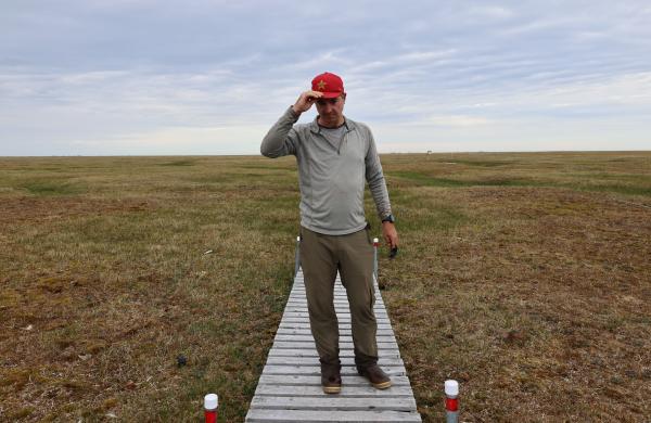 Bob Bolton standing on an elevated wooden pathway over a thawed tundra adjusting his baseball cap.