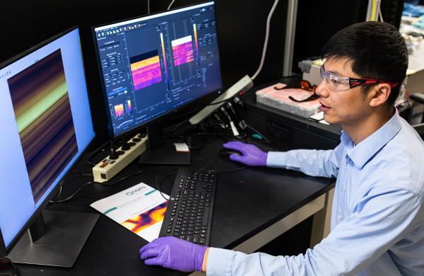 Researcher, on the right, is looking at two computer screens with purple gloves on