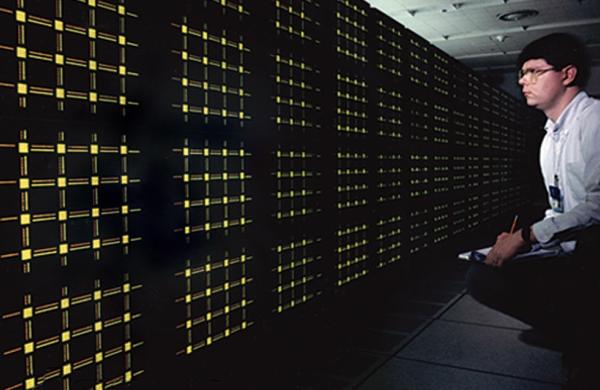 Scientists, right, is kneeling down low to look at a black wall of computer screens with yellow dots in clusters of 12