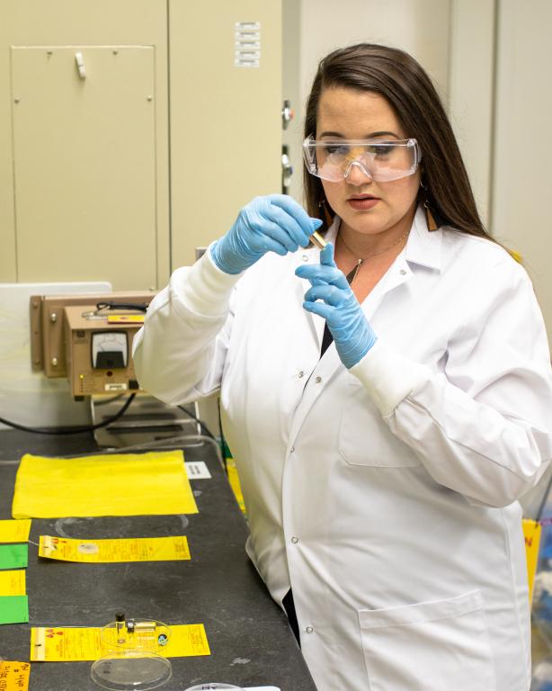 A woman in a lab coat and safety glasses looking at a small material sample.