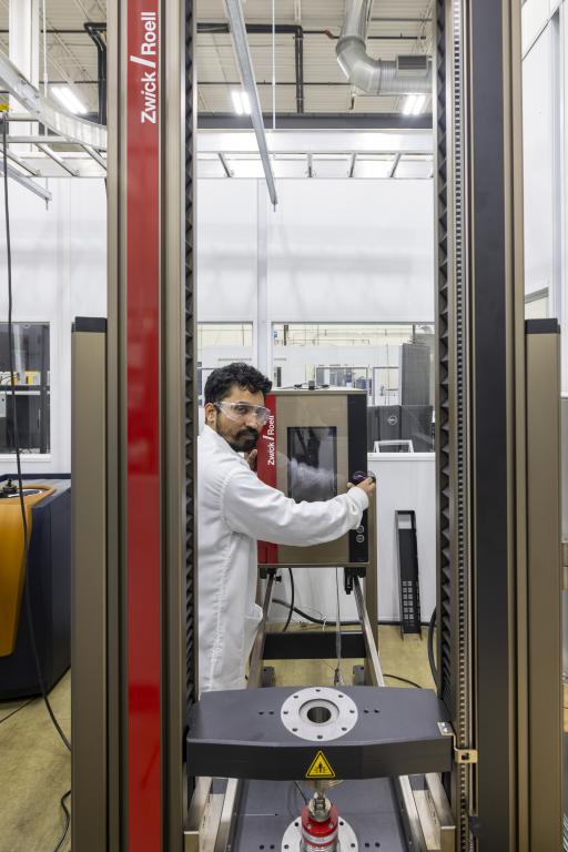 Researcher in lab coat operates mechanical battery testing machine which looks like a tall metal frame with a test platform in the center