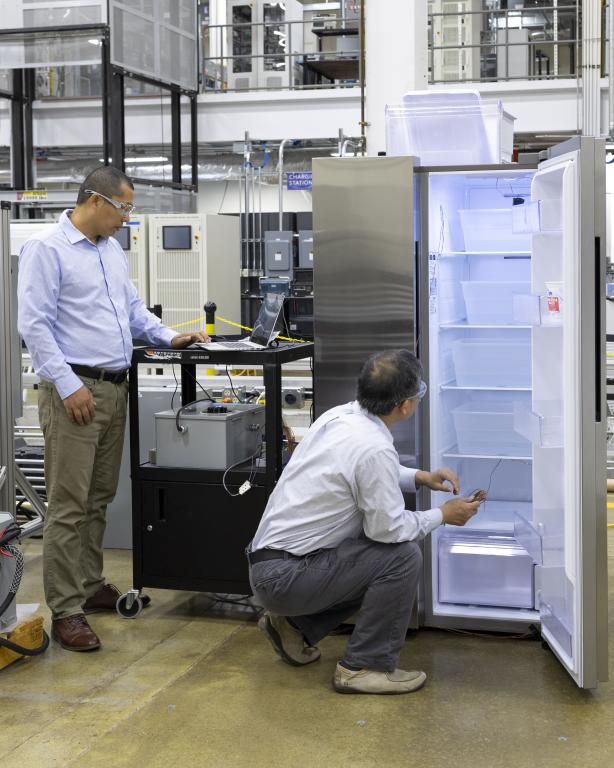 Two men looking at the inside of a silver fridge, one is standing to the left and the other is crouched down on the floor. They are both in business pants and shirts.
