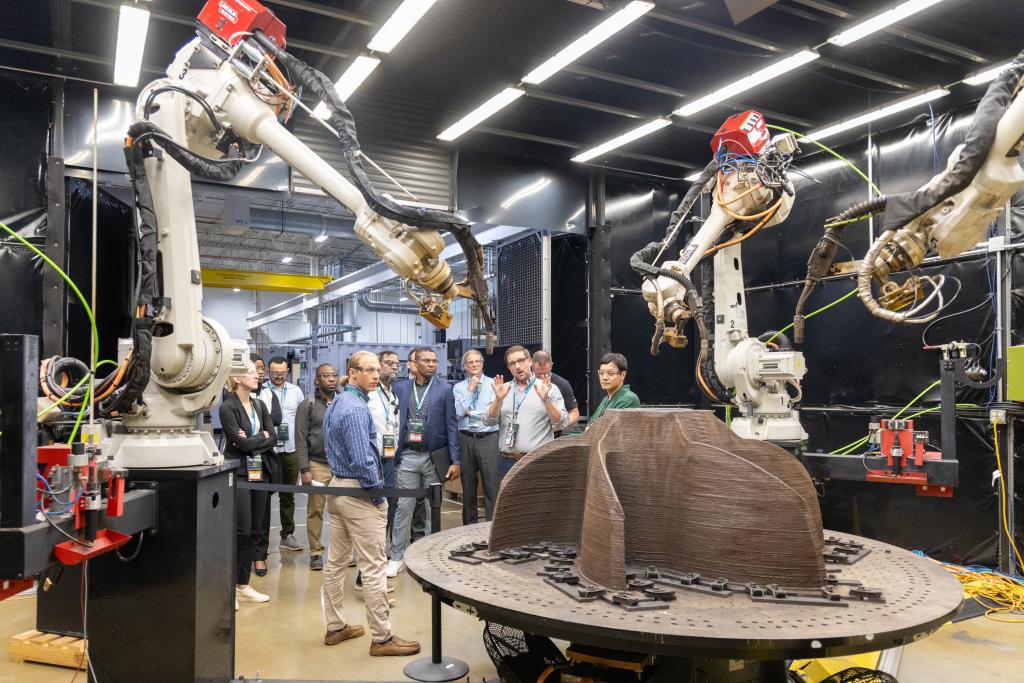 Two robotic arms, one on the left side of the frame and one on the right - are pointing at a brown three pronged shape on a table. There are people in the background looking at this setup.