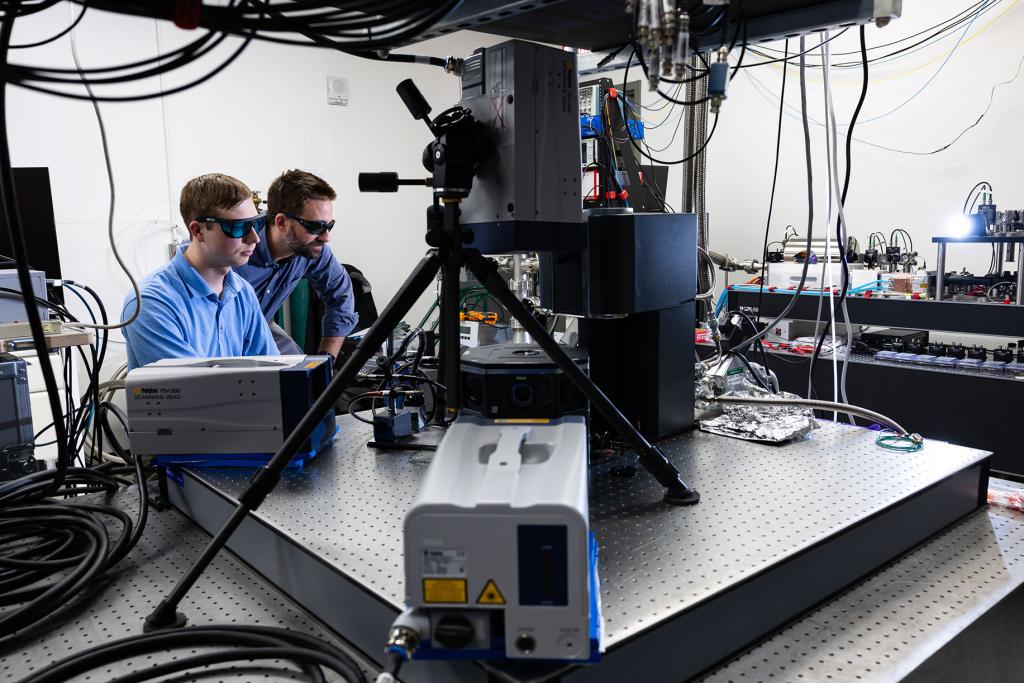 Trevor Michelson and Chris Seck sit at setup in Seck's lab, where the cryogenic ion trap apparatus, center, is surrounded by three heads of the laser scanning vibrometer, each fixed on a different point. The vibrometer will help Seck quantify vibration on the apparatus. Carlos Jones/ORNL, U.S. Dept. of Energy