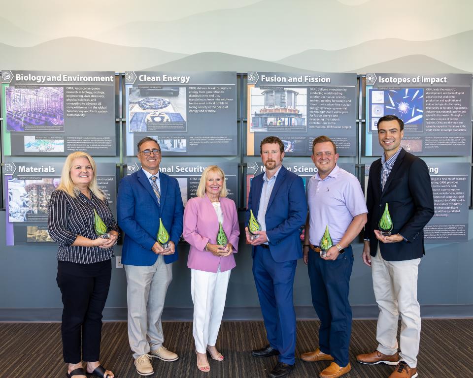 Six people are standing, holding awards, posing in front of a wall with mountain wallpaper. Left to right it goes girl, boy, girl, boy, boy, boy.