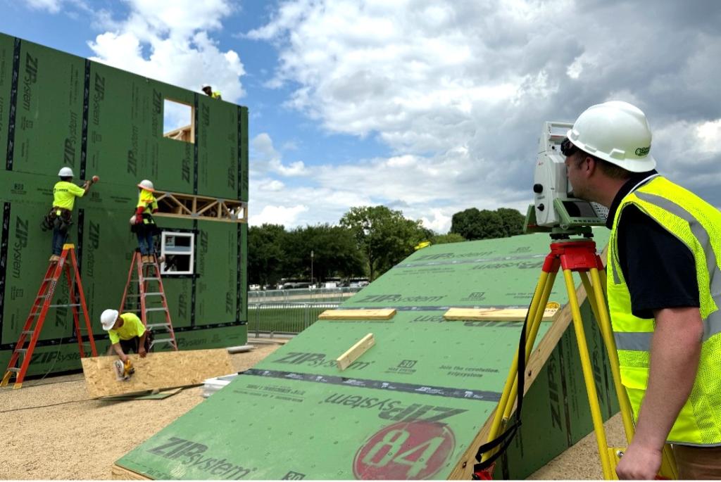Man in bright yellow safety vest and hard hat is looking through a small machine that is pointed at a house being constructed.