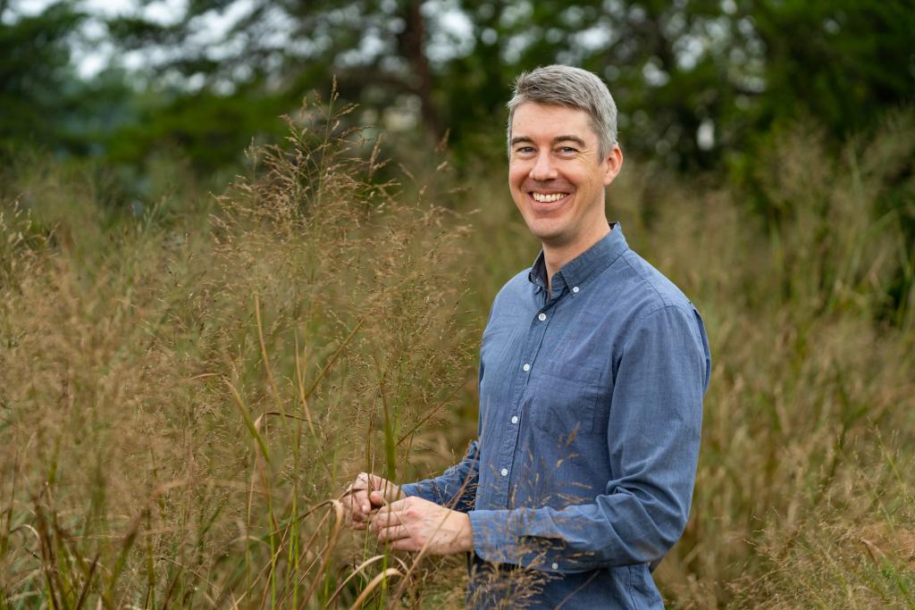 Man in blue button down shirt with short grey hair stands in a field smiling