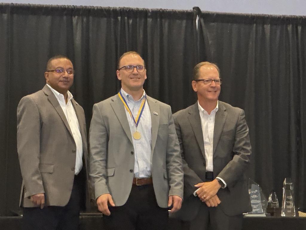 Three men standing in grey suites; the one in the middle had a medal around their neck.