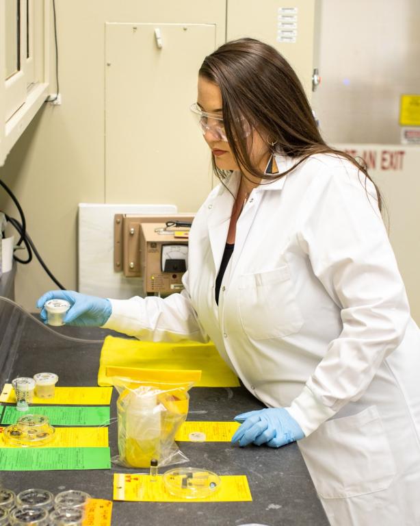 Woman with her hair down is in a lab wearing a white coat and protective safety glasses.