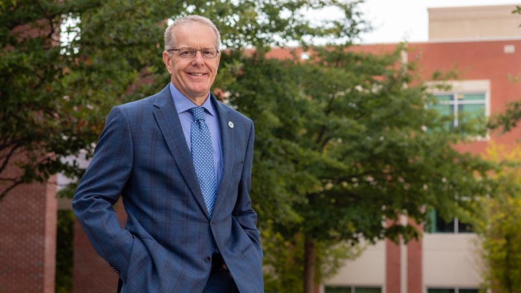 Man in blue suit and tie with glasses and grey hair poses outside in front of trees and a brick building