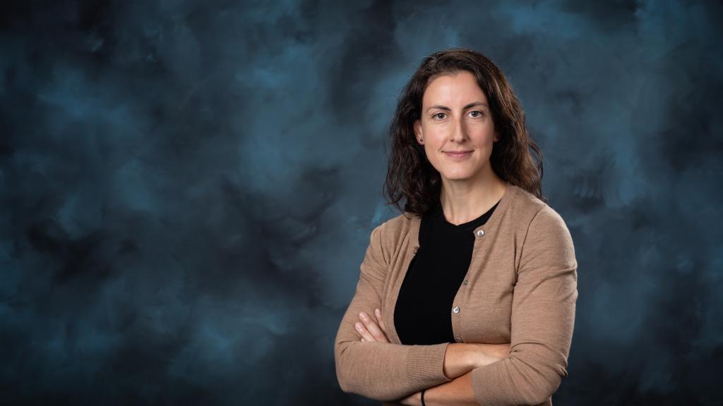 Image is of woman with dark hair crossing her arms and posting for a photo in front of a blue backdrop. She is wearing a black shirt with a tan cardigan.
