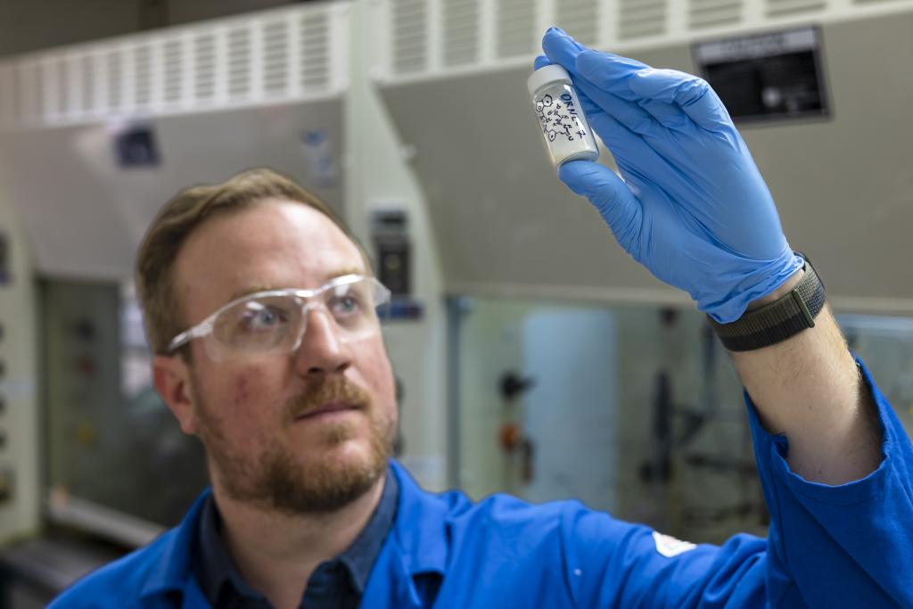 Man in blue lab coat and gloves looks at a sample, holding it up to the light.