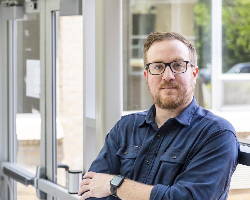 Man is leaning against the window, arms crossed in a dark navy button up.